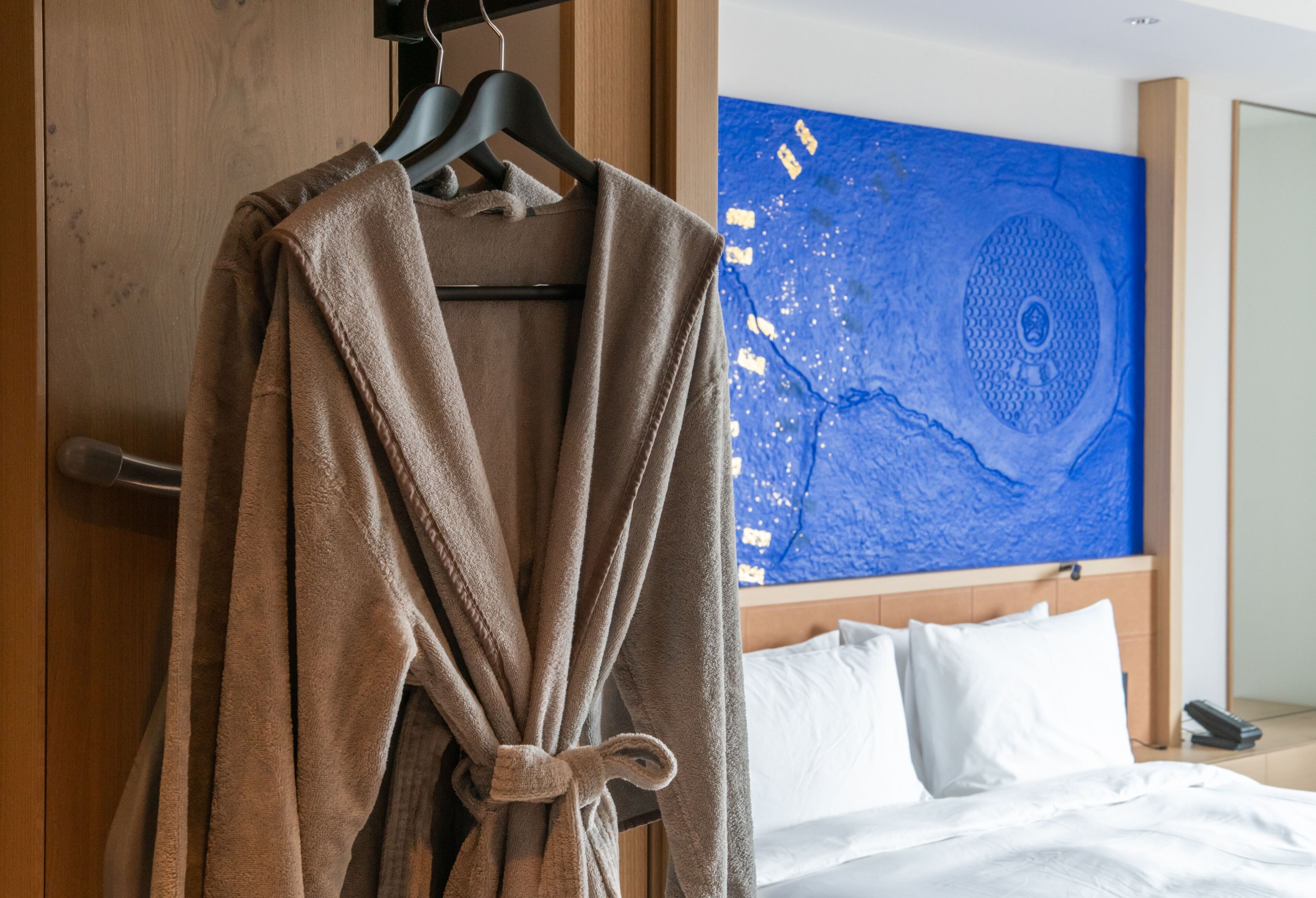 A hotel room with a bed, a robe hanging on a rack, and a large blue artwork on the wall.