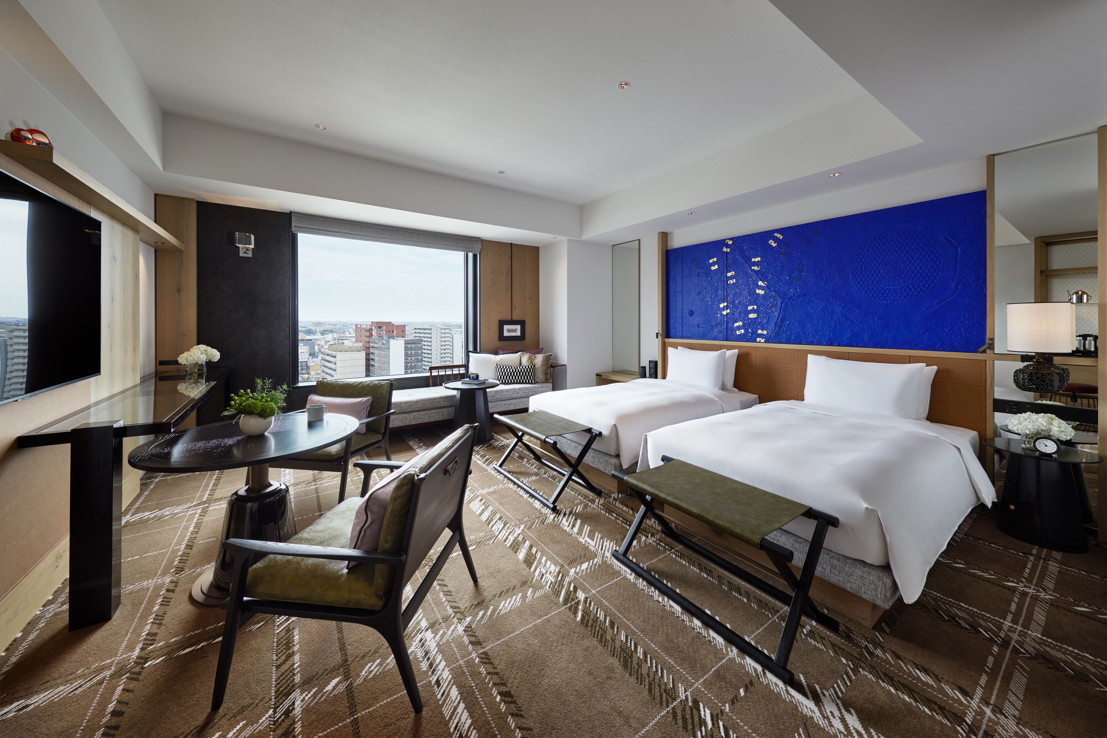 A hotel room with two beds, a desk, a chair, a small table, a large window, and a city view.