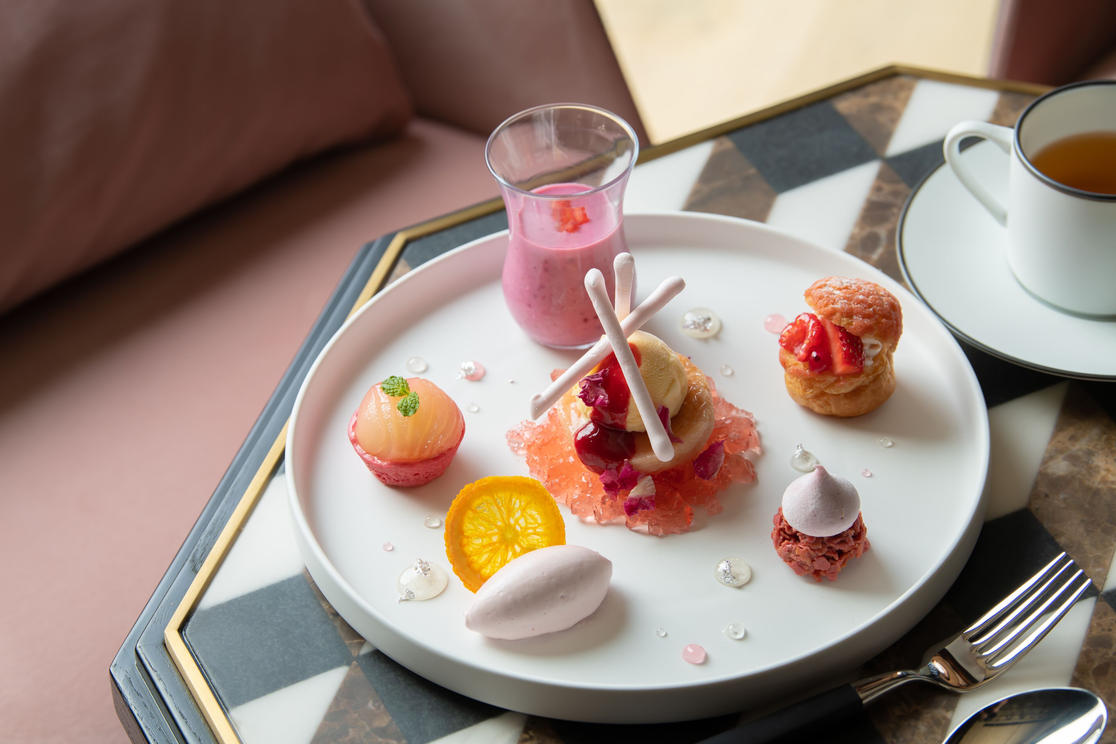 A dessert plate with various sweet treats and a glass of a pink beverage, accompanied by a cup of tea on a table.
