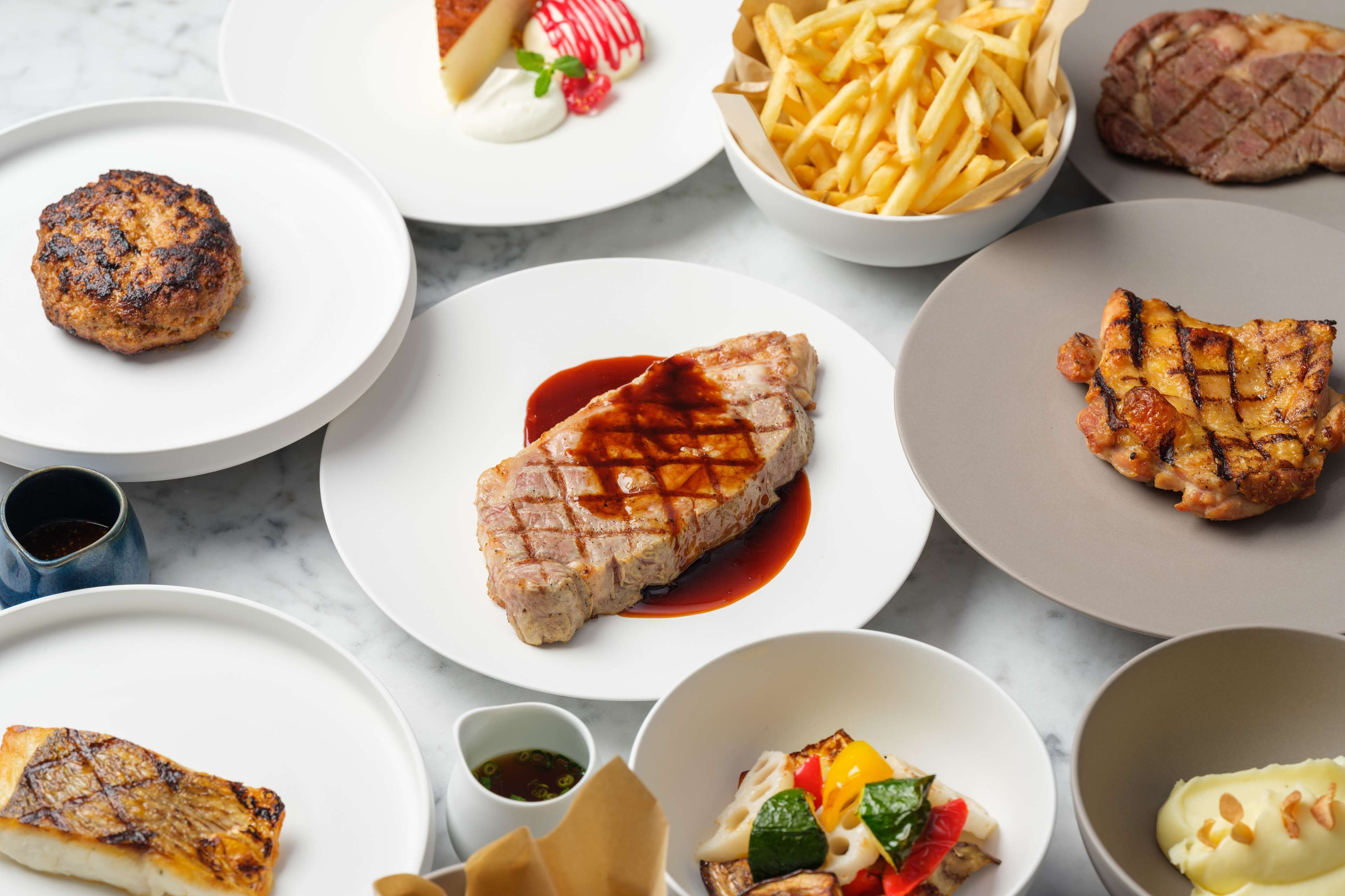 A variety of grilled meats and sides on plates, including steak, chicken, and a burger.
