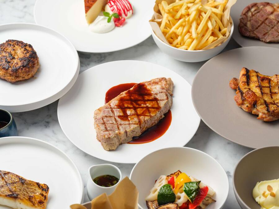 A variety of grilled meats and sides on plates, including steak, chicken, and a burger.