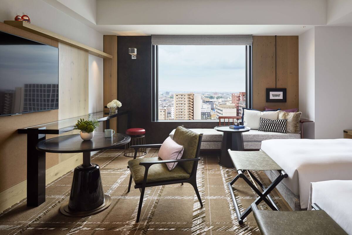 Ein Hotelzimmer mit großem Fenster, einem Bett, einem Sofa, einem Esstisch und einem kleinen Tisch mit Pflanze.