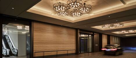 A modern hotel lobby with a spacious layout, a central chandelier, and an escalator leading to another floor.