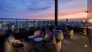 A rooftop lounge with wicker furniture, round tables, and a city view at dusk.