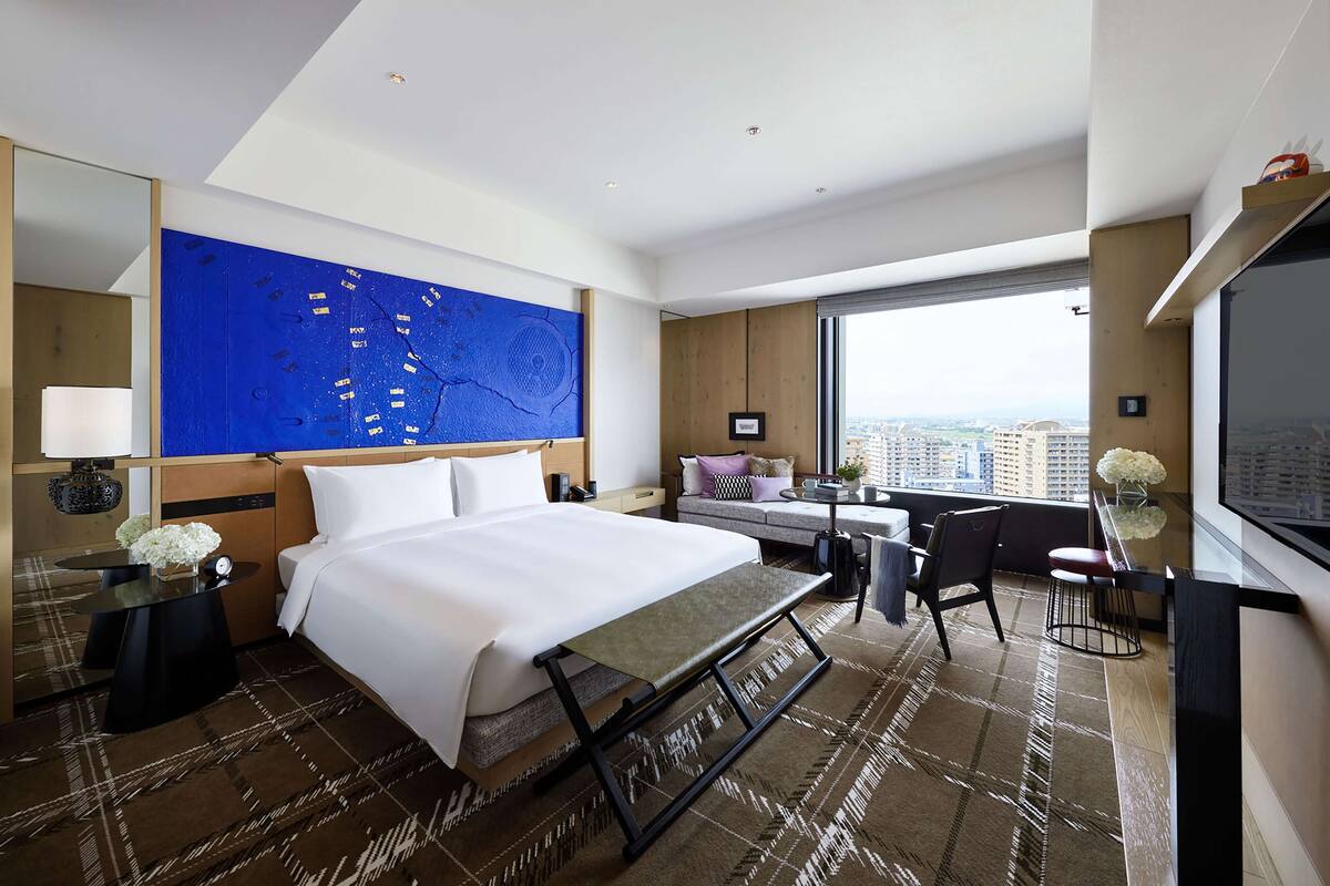 Ein Hotelzimmer mit einem großen Bett, einem Schreibtisch, einem Sessel, einem Fernseher und Blick auf die Stadt.