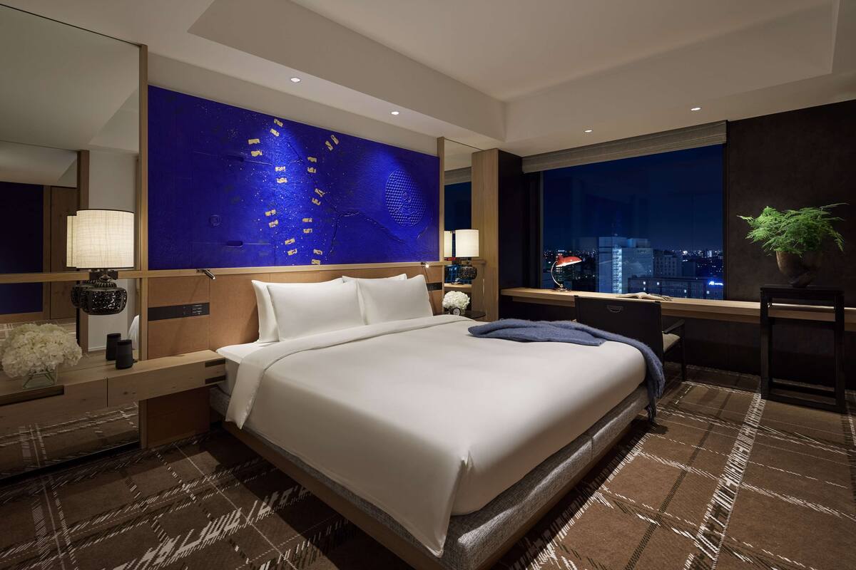 Ein modernes Hotelzimmer mit einem großen Bett, einem Schreibtisch, Blick auf die beleuchtete Stadt und einer blauen Wand als Dekoration.