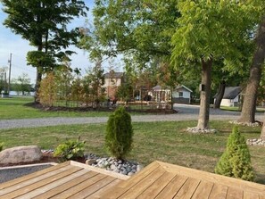Una terraza de madera con borde de piedra, rodeada de césped, arbustos pequeños y algunos árboles.