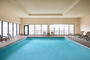 A large indoor swimming pool with clear views of the city through large windows, surrounded by lounge chairs and a small table.