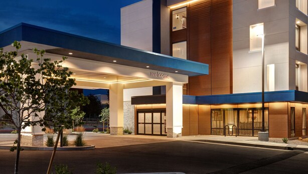 A modern hotel entrance with a 'Welcome' sign, a covered driveway, and a landscaped area with trees and shrubs.