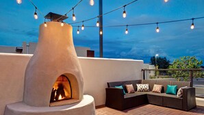 A modern outdoor seating area with a stone-built fireplace, string lights, and a cushioned sofa.