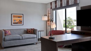 A modern hotel room with a grey sofa, a small table, a desk, a TV, and a large window offering a city view.