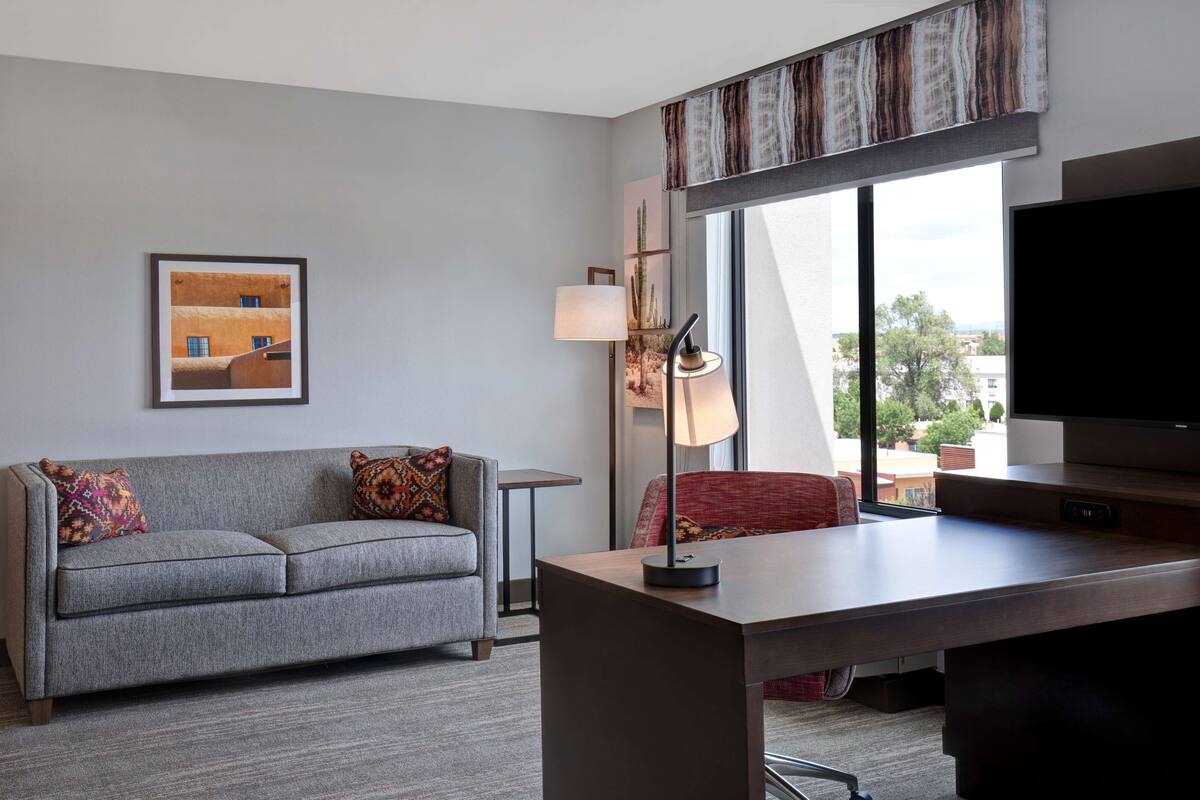 Une chambre d’hôtel moderne avec un canapé gris, une petite table, un bureau, un téléviseur et une grande fenêtre donnant sur la ville.