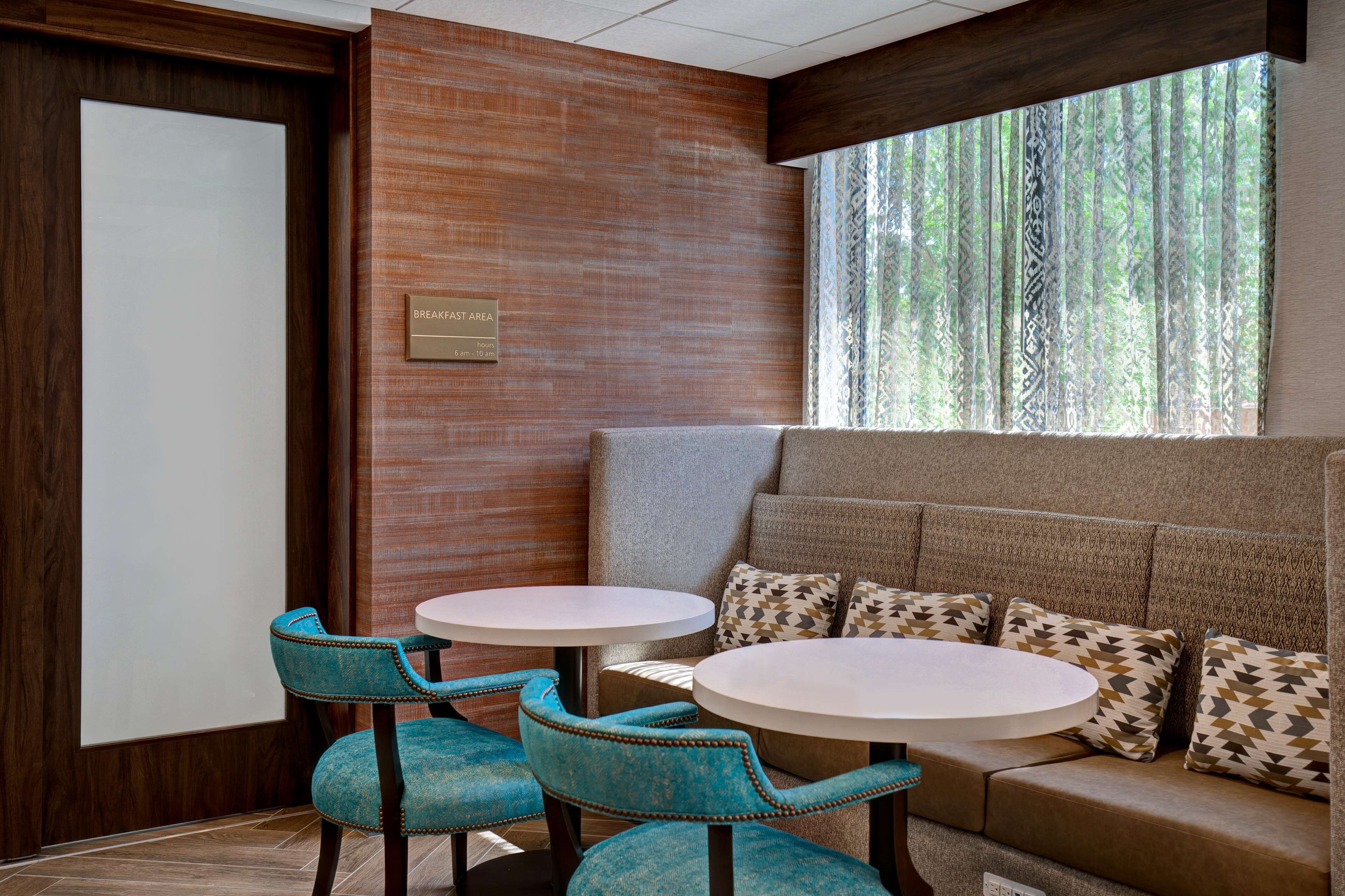 A break area with two round tables, teal upholstered chairs, a cushioned bench with patterned pillows, and a view of trees through patterned curtains.