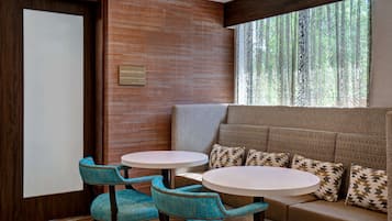 A break area with two round tables, teal upholstered chairs, a cushioned bench with patterned pillows, and a view of trees through patterned curtains.