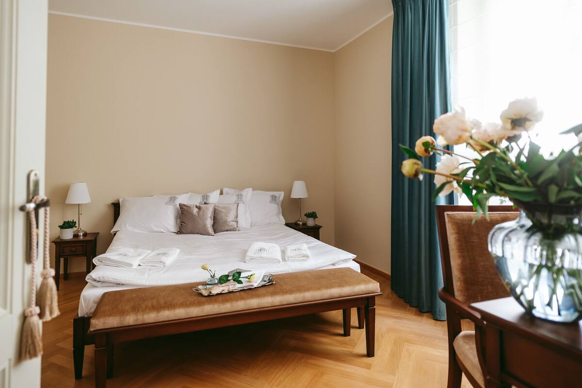 Ein ordentlich bezogenes Bett mit weißen Leinen und Kissen, ein Nachttisch mit Lampe, eine Holzbank und eine Vase mit Blumen.