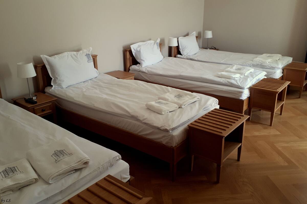 Ein Hotelzimmer mit drei Einzelbetten, jedes mit einem hölzernen Kopfteil, weißer Bettwäsche und einem Handtuch mit einem Gebäude-Design. Es gibt Nachttische mit Lampen und eine kleine Schublade.