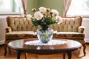 A round wooden table with a clear glass vase containing white flowers, placed on a lace doily. In the background, there is a patterned sofa with a cushion.