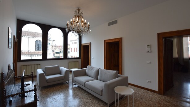 A spacious living room with a large window, a chandelier, a sofa, a coffee table, and a wooden side table.