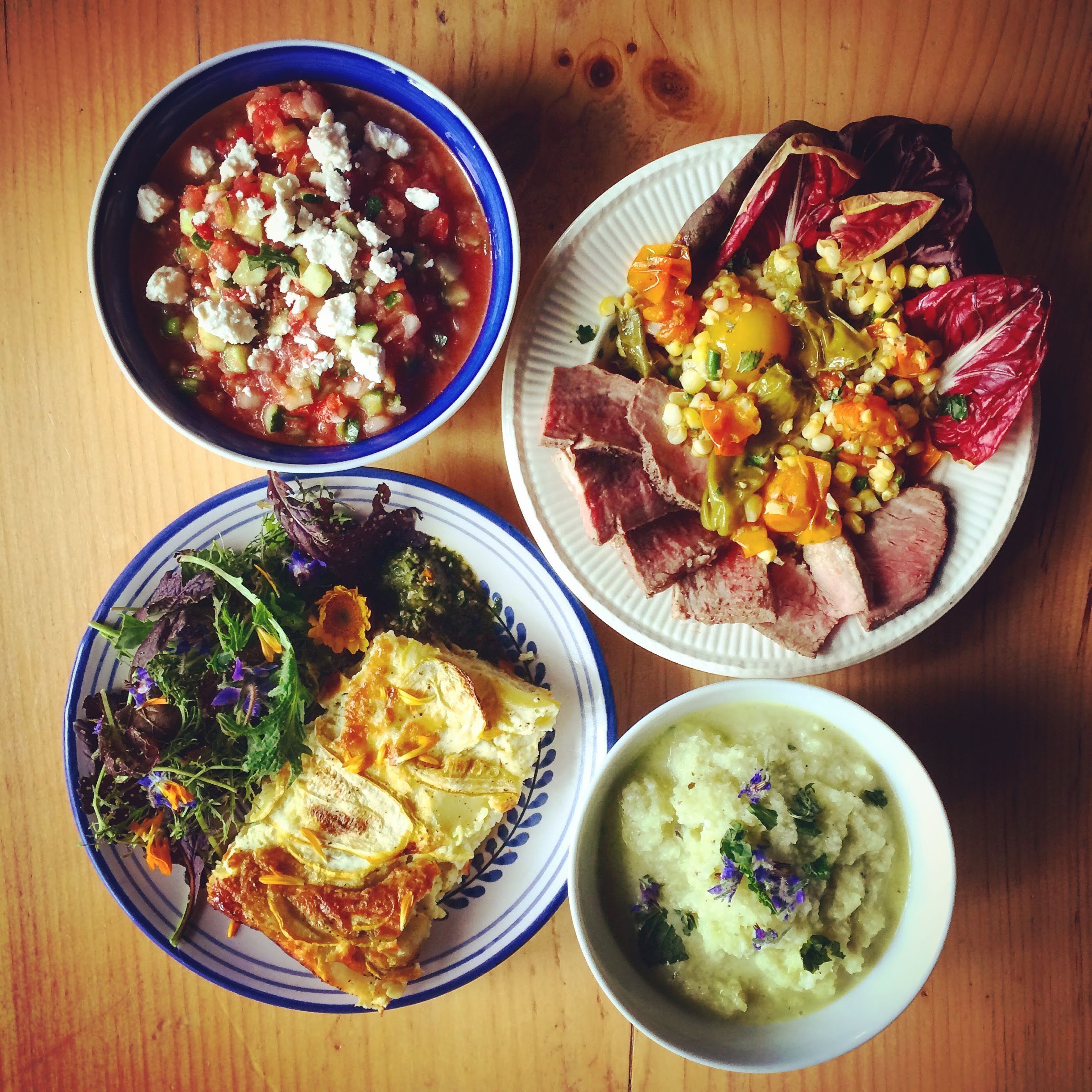 Una mesa con cuatro platos: un tazón de sopa con verduras y queso, un plato con carne y verduras, una ensalada de hojas verdes y flores, y un tazón de puré de patatas con hierbas.