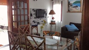A living room with a glass coffee table, a sofa, a TV mounted on a wall with newspaper clippings, and a painting of a beach. - House for rent in the Center of Ubatuba (Ubatuba)