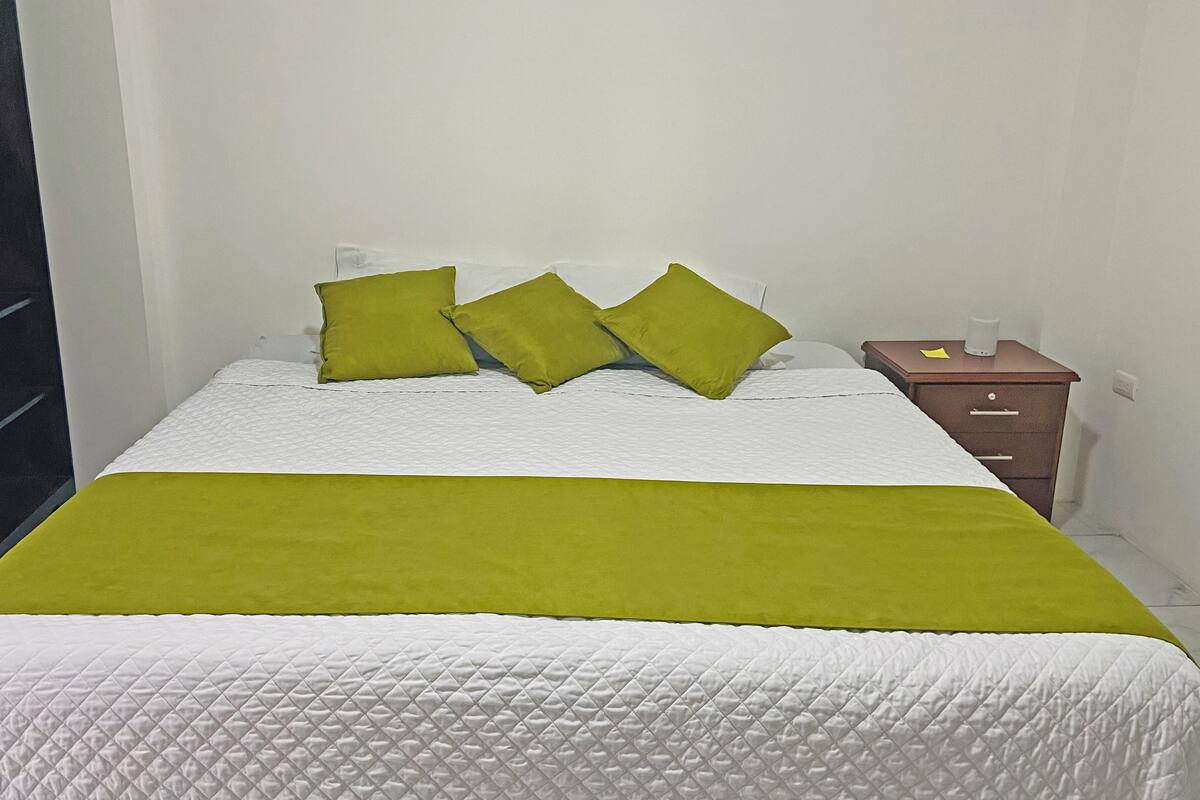 A bed with a white quilted mattress and a green throw blanket. There are three green pillows on the bed. A wooden nightstand with a white lamp and a small drawer is to the right of the bed.