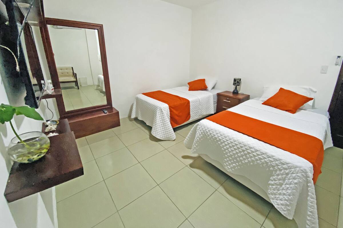 A hotel room with two beds, each with white bedding and orange accents, a large mirror, a small table with a plant, and a tiled floor.