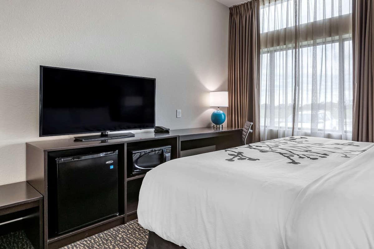 A hotel room with a flat-screen TV, a small refrigerator, a microwave, a desk with a lamp, and a bed with a white bedspread.