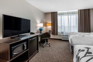 A hotel room with a flat-screen TV, a desk with a lamp, a chair, a bed, and a large window with curtains.