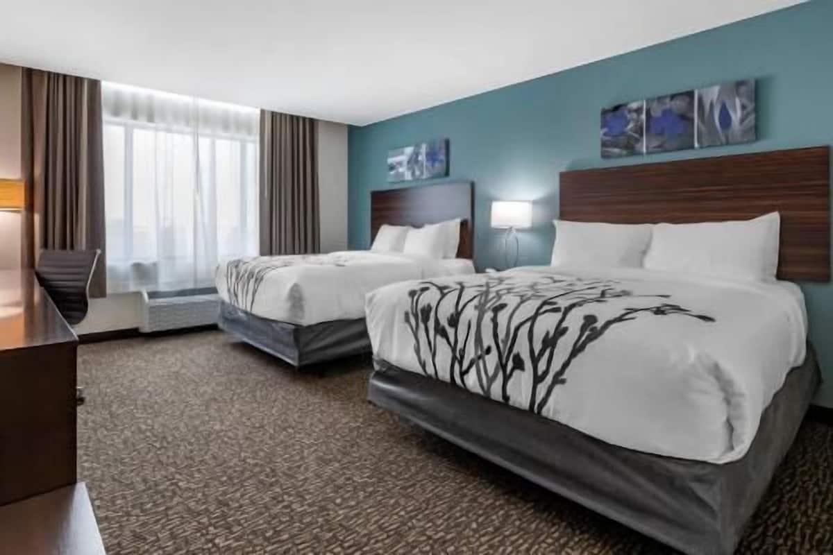 A hotel room with two beds, a desk, a chair, a lamp, and artwork on the wall.