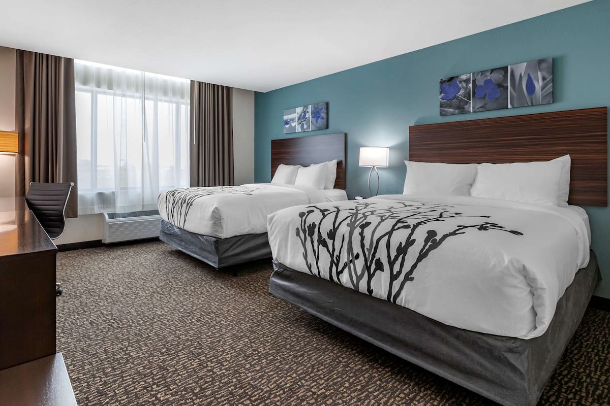 A hotel room with two beds, a desk, a chair, a window with curtains, and artwork on the walls.