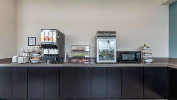 A self-service breakfast bar with a coffee machine, refrigerator, microwave, and various food items.