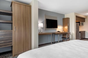 Habitación de hotel con cama, escritorio con televisor de pantalla plana, lámpara, silla y kitchenette con microondas y refrigerador.