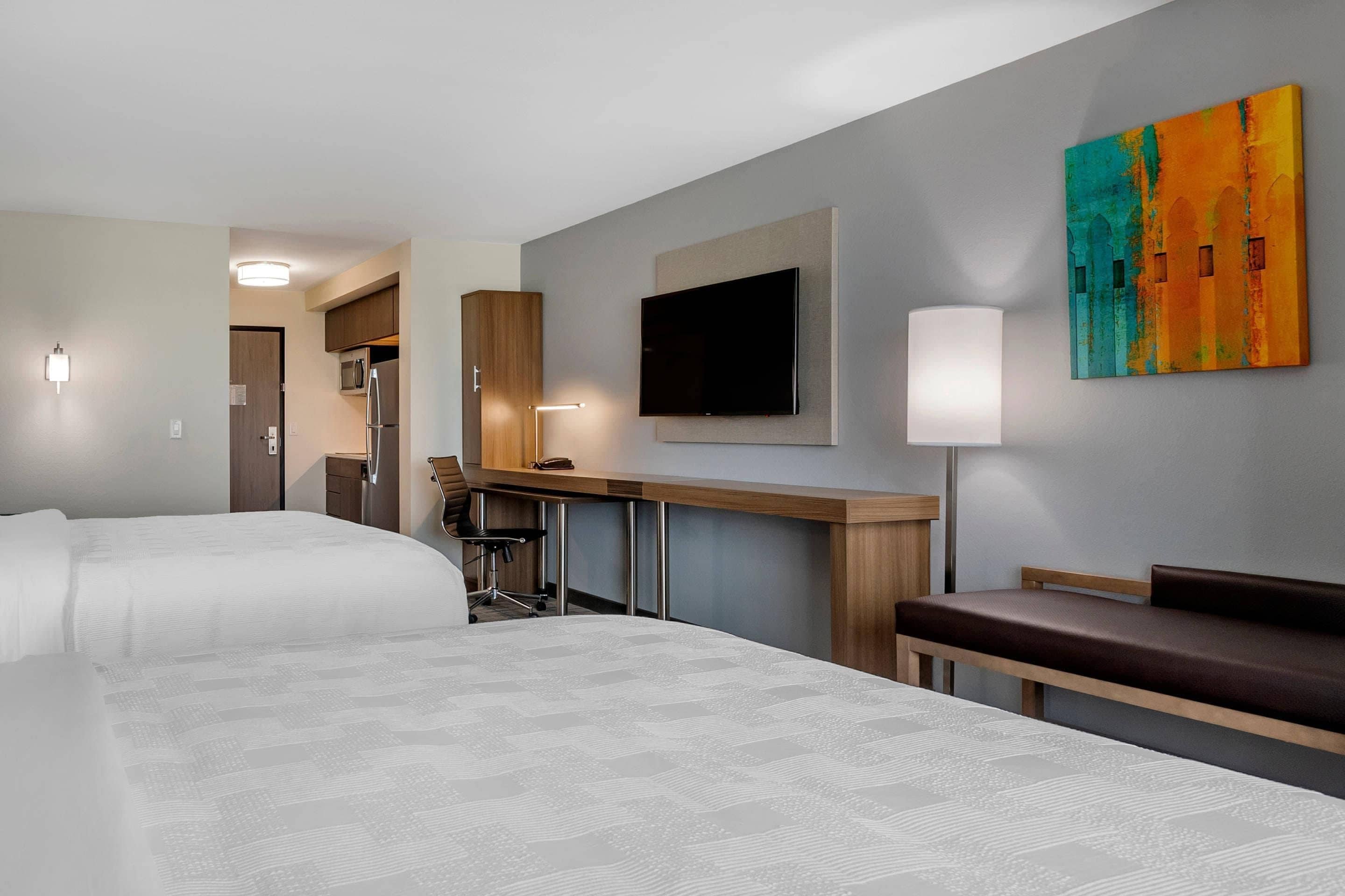 A hotel room with two beds, a desk with a chair, a flat-screen TV, a painting, and a lamp.