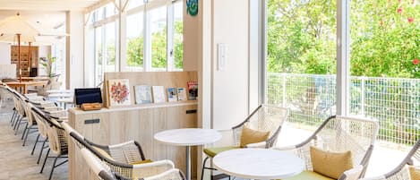 A modern indoor seating area with wicker chairs and round tables, a wooden counter with a computer, and large windows offering a view of greenery.