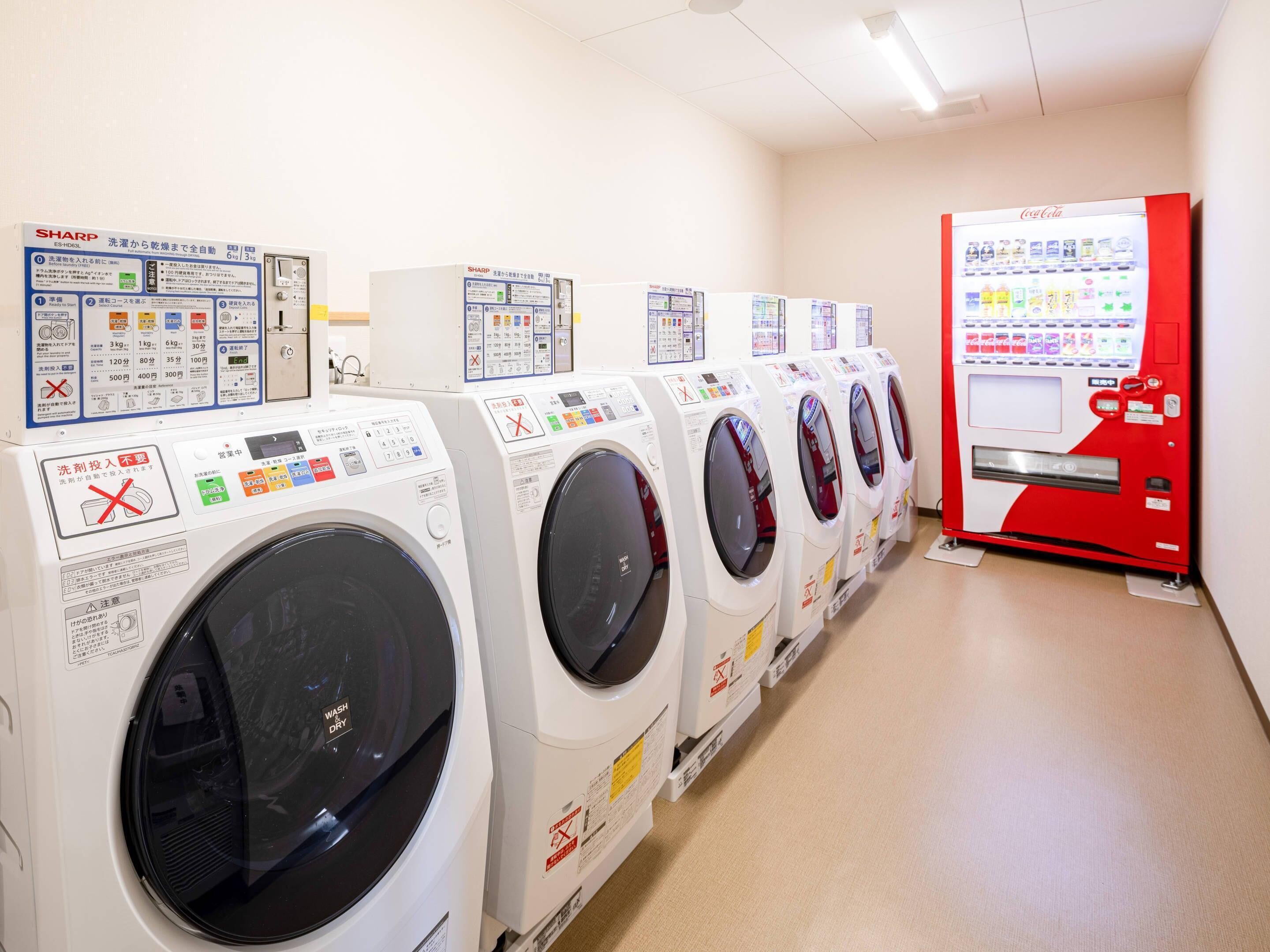 A row of front-loading washing machines with control panels, a Coca-Cola vending machine, and a wall-mounted Sharp washing machine.
