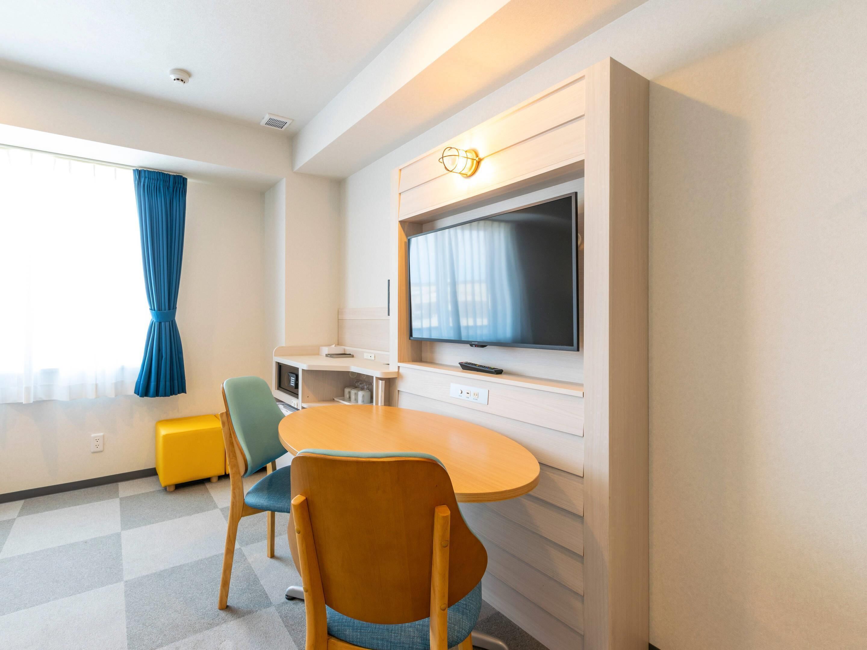 A hotel room with a TV mounted on the wall, a wooden table, two chairs, a small kitchenette, and a window with curtains.