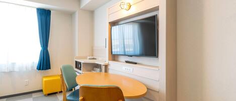 A hotel room with a TV mounted on the wall, a wooden table, two chairs, a small kitchenette, and a window with curtains.
