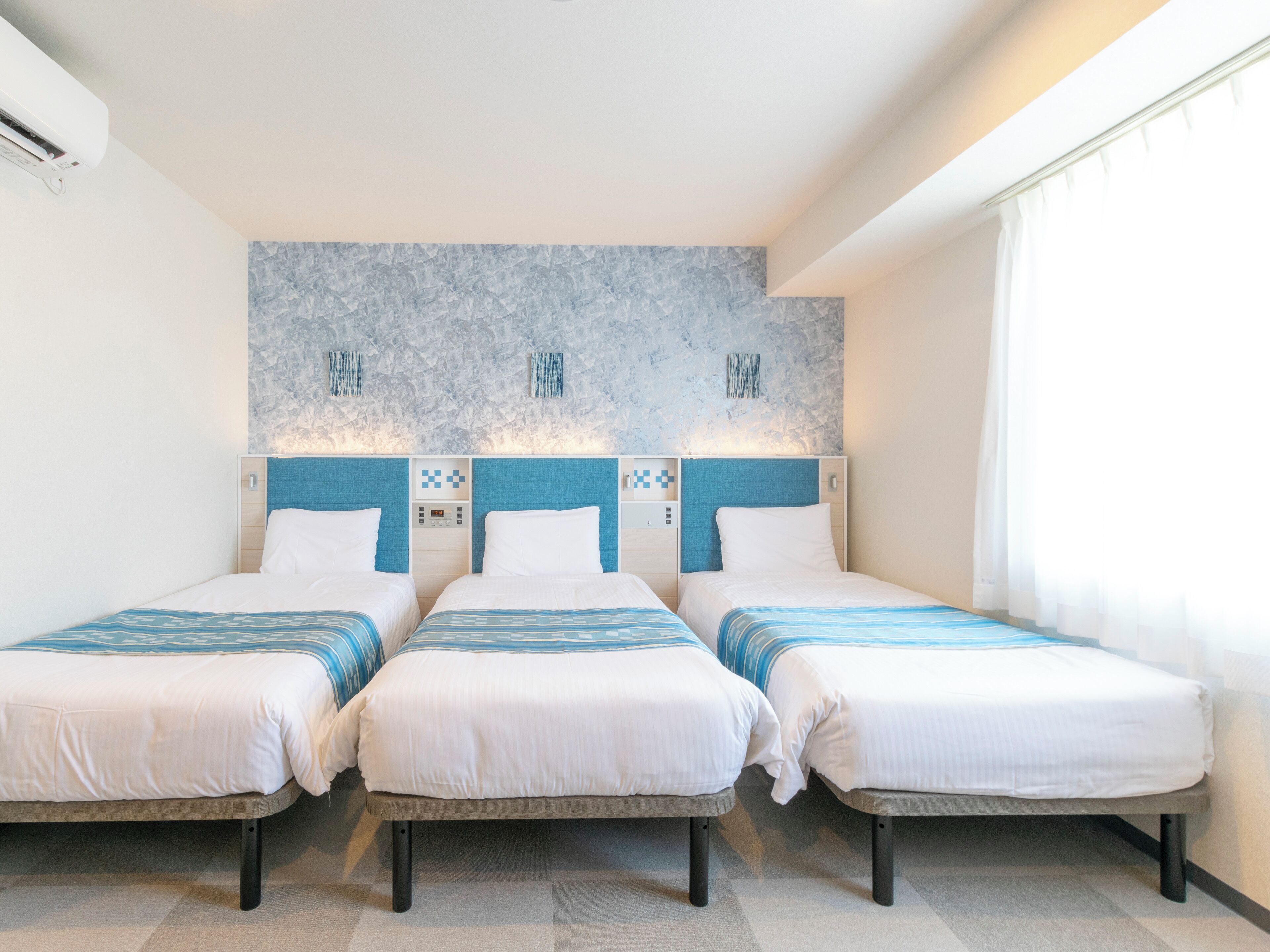 A hotel room with three single beds, a marble-patterned headboard, a control panel, and a window with curtains.