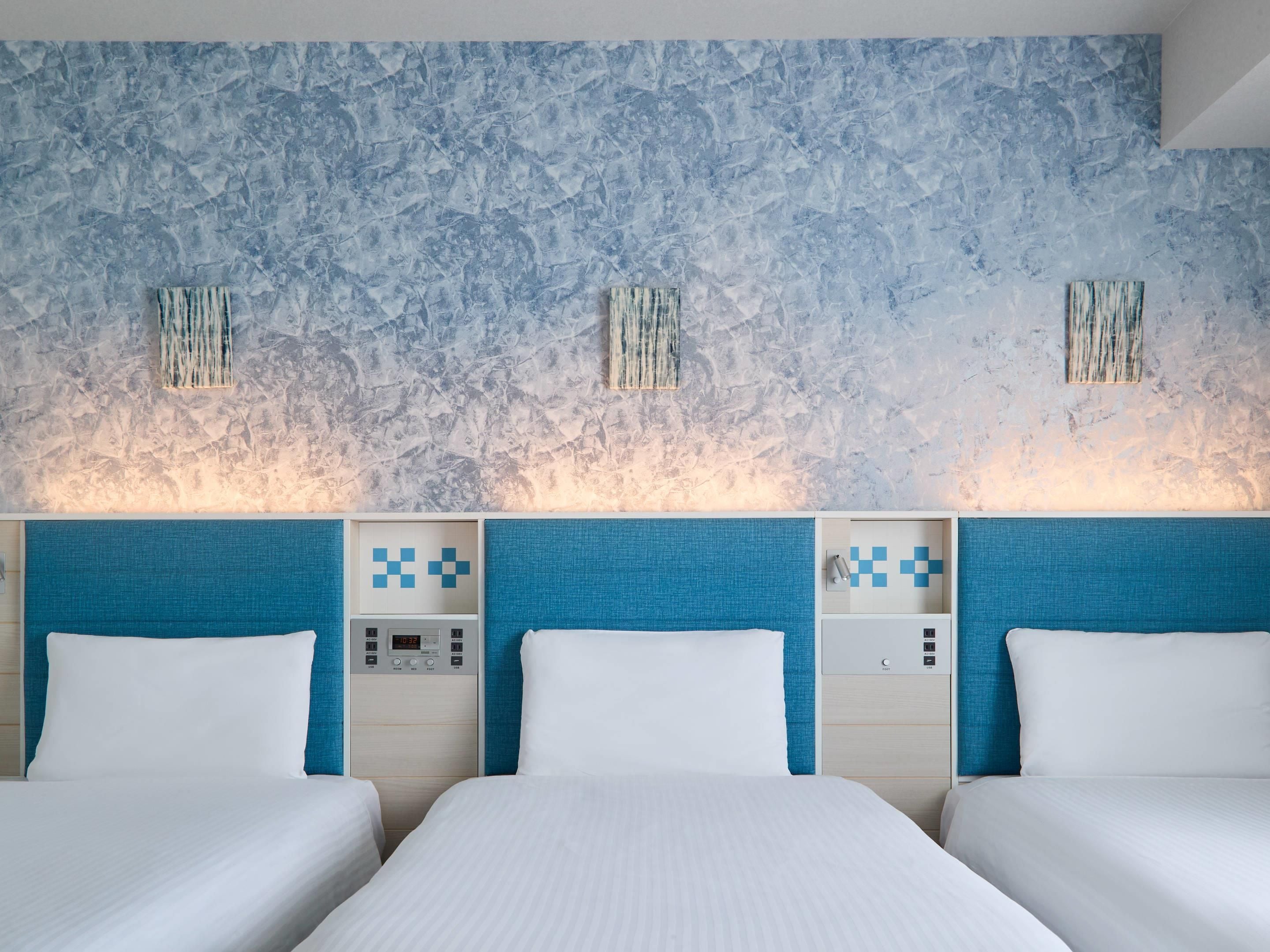 A hotel room with a textured blue wall, a bed with white bedding, and a control panel with a digital clock.