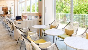 Un área de estar interior moderna con sillas de mimbre y mesas redondas, una barra de madera con una computadora, y ventanas amplias con vista a zonas verdes.