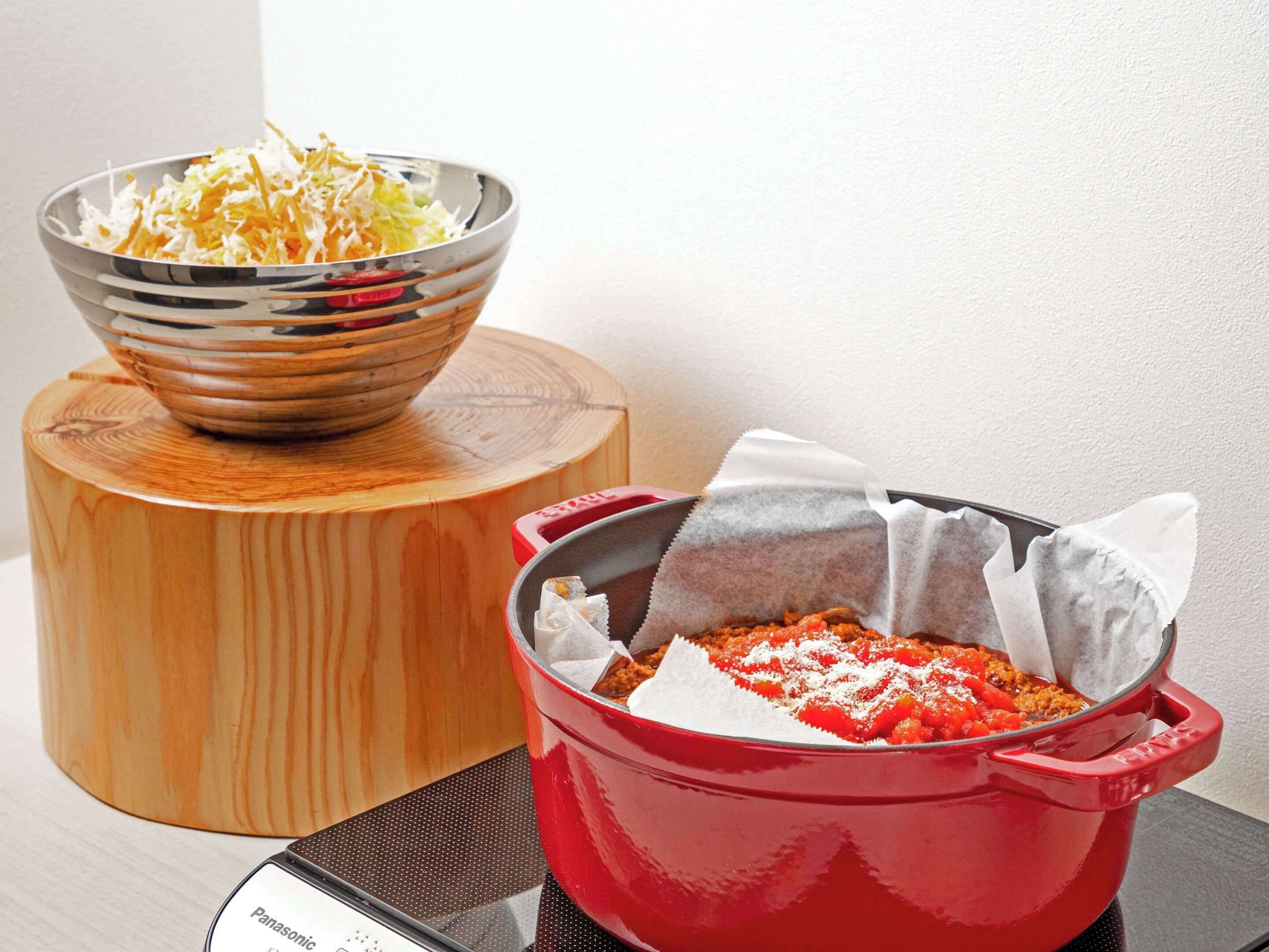 A red pot on a stove with a white label, a bowl of shredded vegetables, and a wooden cutting board.