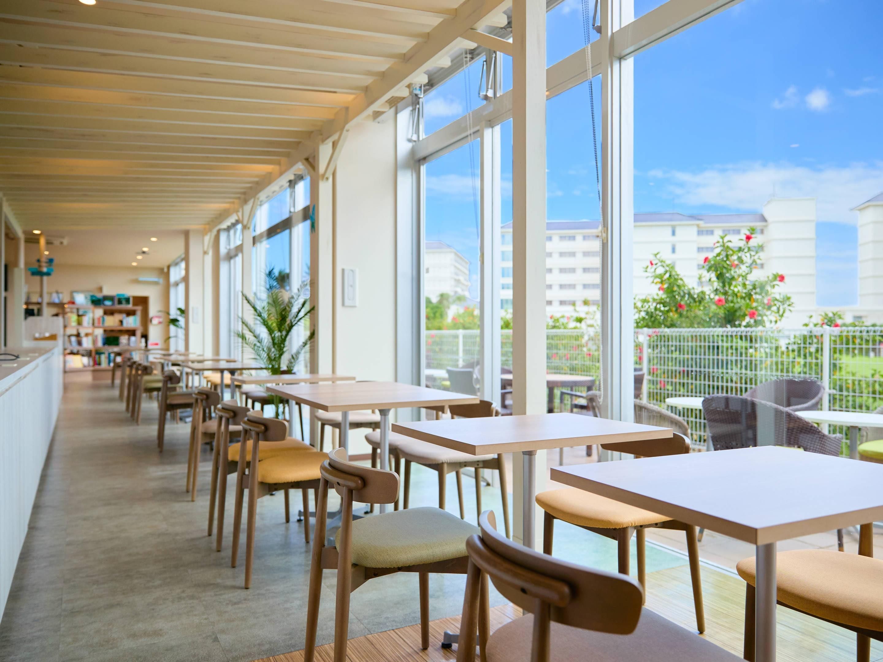 A spacious indoor area with tables and chairs, large windows, and a view of buildings and greenery outside.