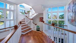 Un escalier spacieux, doté de rampes en bois et de balustrades blanches, mène à un palier avec de grandes fenêtres offrant une vue sur la verdure.