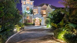 Une grande maison à plusieurs étages, dotée d’une tour imposante, entourée d’une végétation tropicale luxuriante et d’une allée bien éclairée.