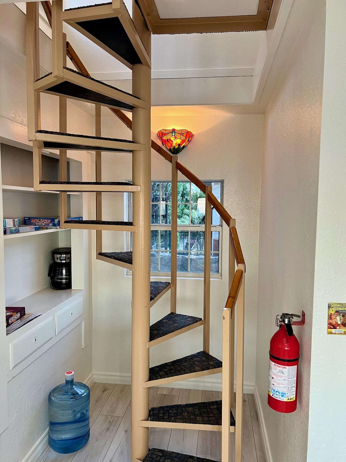A spiral staircase with wooden railings and carpeted steps, a fire extinguisher on the wall, and a water dispenser on the floor.
