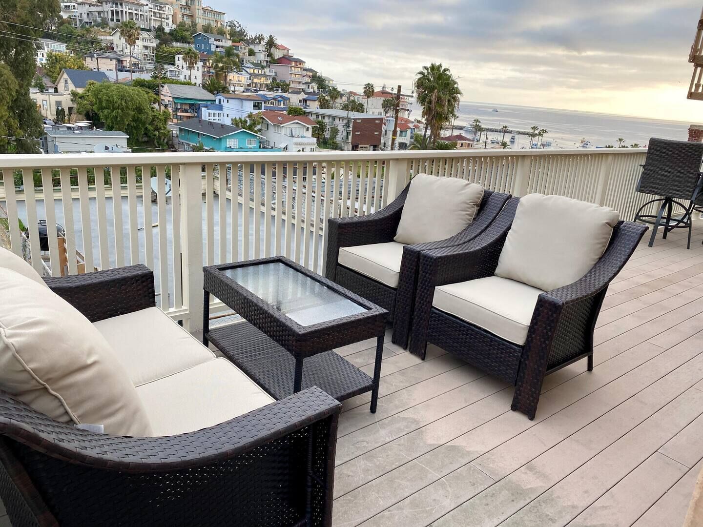 A balcony with wicker furniture, a glass table, and a view of the ocean and hillside buildings.