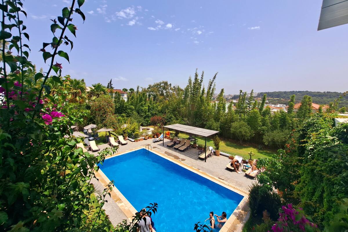 A swimming pool with lounge chairs and a covered seating area, surrounded by lush greenery and a view of a distant town.
