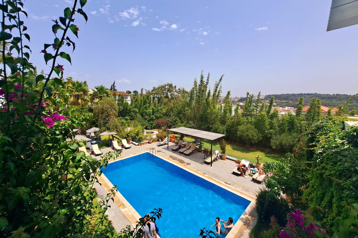 Una piscina con sillones de descanso y una zona de estar cubierta, rodeada de exuberante vegetación y vistas a un pueblo lejano.