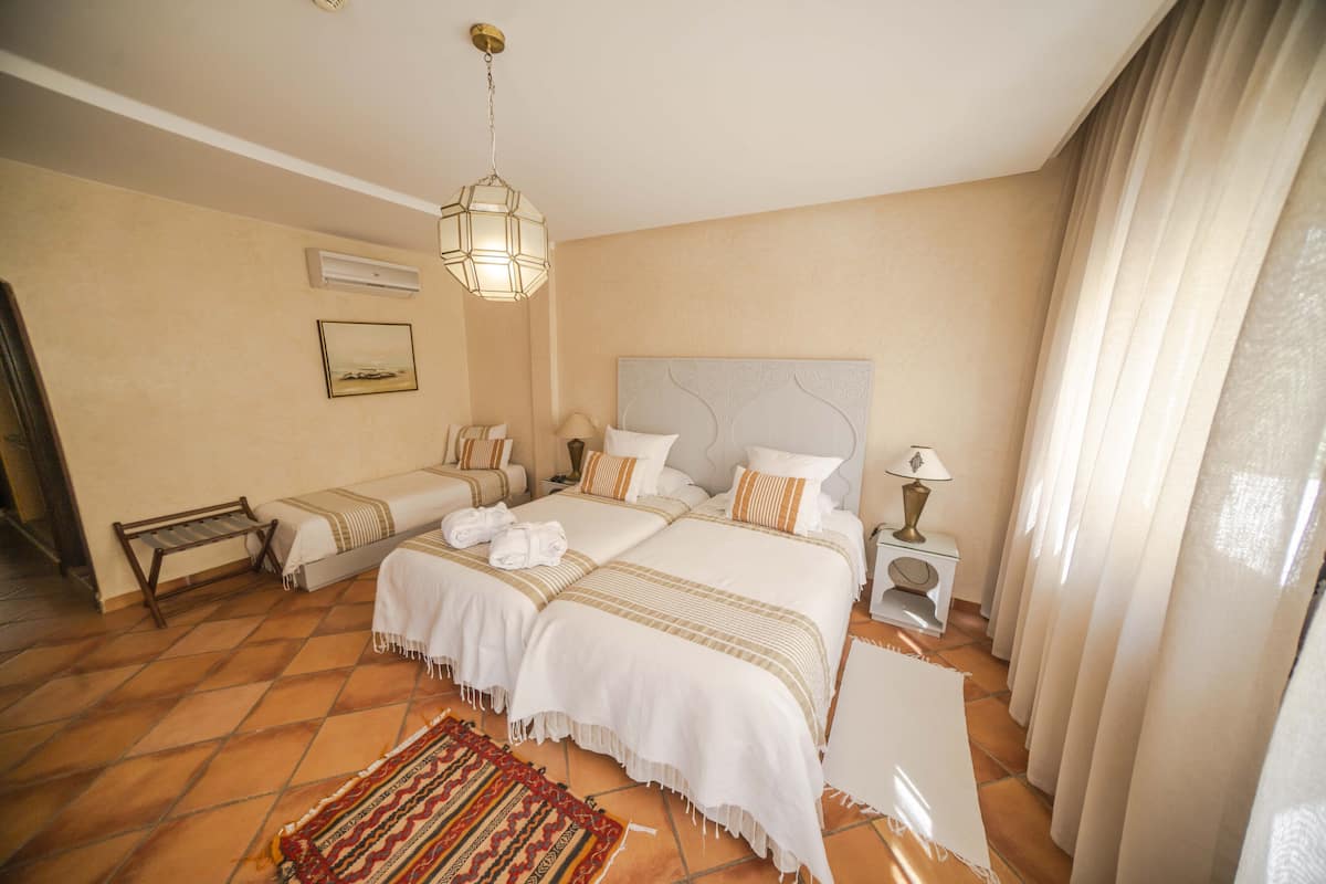 A hotel room with two beds, a bench, a bedside table with a lamp, and a large window with curtains.