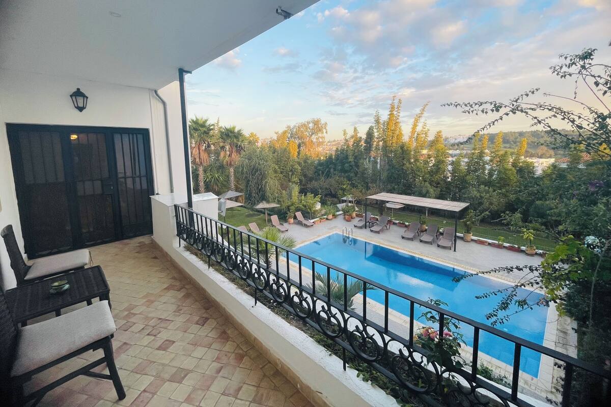 A balcony with a view of a swimming pool, outdoor furniture, and a garden.